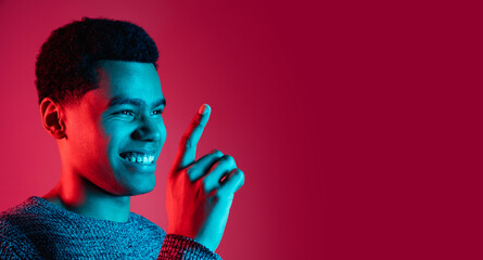 Pointing. African-american man's portrait on red background in multicolored neon light. Beautiful...