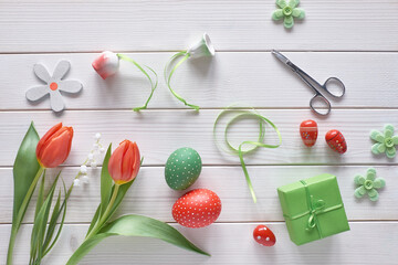 Fototapeta na wymiar Flat lay with red tulips in rattan wreath basket and green grass. Decorative painted Easter eggs. Springtime, Easter background on rustic off white wooden table. Gift box, ribbon and scissors.