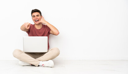 Teenager man sitting on the flor with his laptop making phone gesture and pointing front