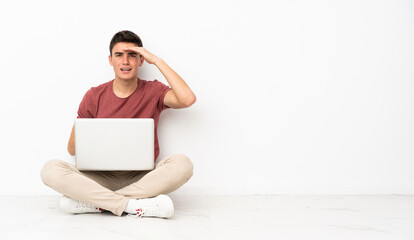 Teenager man sitting on the flor with his laptop looking far away with hand to look something