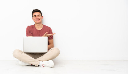 Teenager man sitting on the flor with his laptop presenting an idea while looking smiling towards