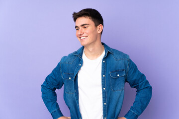 Teenager caucasian  handsome man isolated on purple background posing with arms at hip and smiling