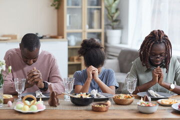 Portrait of modern African-American family saying grace at dinner table while celebrating easter at...