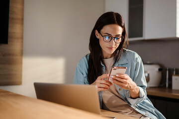 Serious charming woman using smartphone while working with laptop - Powered by Adobe