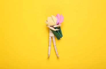 Wooden puppet holds origami tulips on a yellow background. Top view