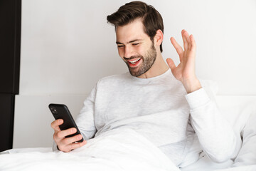 Smiling man waving hand and using mobile phone while resting in bed