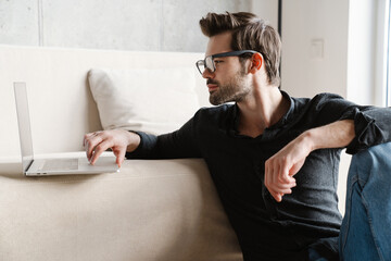 Fototapeta premium Focused unshaven man using laptop while sitting on floor at home