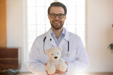 Portrait of smiling young Caucasian male pediatrician in white medical uniform hold fluffy teddy...