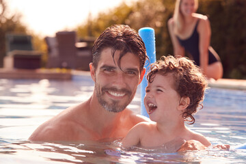 Family In Outdoor Pool On Summer Vacation Teaching Son To Swim With Noodle Swimming Aid