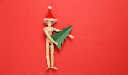 Wooden puppet with santa hat holding paper christmas tree on red background