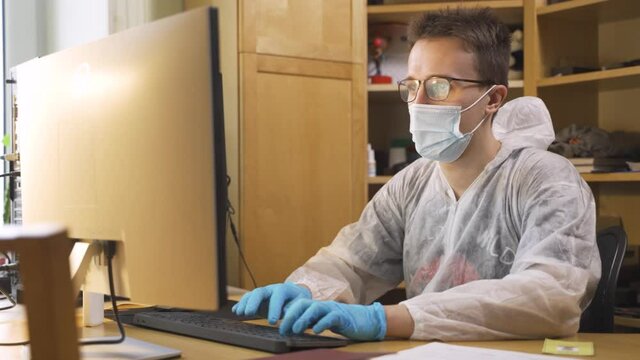 Close up portrait of young handsome Caucasian male doctor sitting in cabinet in mask and face shield work in medical center during quarantine typing on computer and looking at screen Physician concept