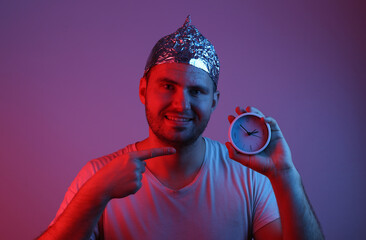 Cheerful Bearded Man in foil hat holds clock in red-blue neon light. Conspiracy
