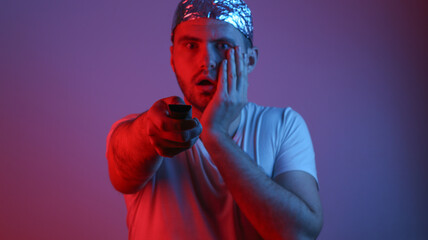 Surprised Bearded Man in a foil hat holds tv remote in red-blue neon light. Conspiracy