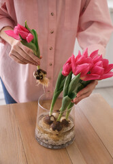 Woman putting pink tulips with bulbs into vase at table indoors, closeup