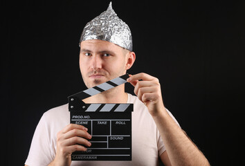 Man in aluminum foil hat holding clapperboard on black background