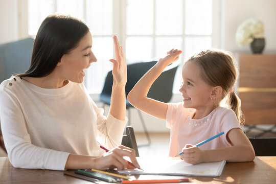Overjoyed young Caucasian mother and small 7s daughter give high five drawing together in album. Happy mom and little girl child celebrate accomplishment involved in funny learning activity painting.