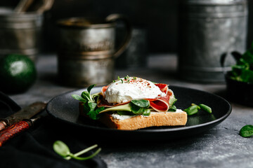 Breakfast. Sandwich with ham, poached egg, avocado and spinach in a plate on a dark background