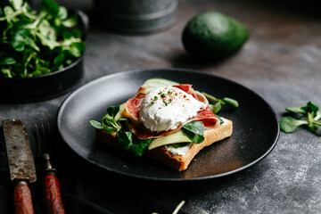 Breakfast. Sandwich with ham, poached egg, avocado and spinach in a plate on a dark background