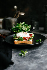 Breakfast. Sandwich with ham, poached egg, avocado and spinach in a plate on a dark background