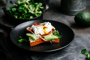 Breakfast. Sandwich with ham, poached egg, avocado and spinach in a plate on a dark background