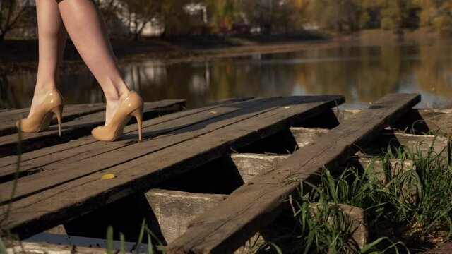 Bare slender legs with veiny feets in beige patent leather heels, climbing up on an old dilapidated platform by the lake