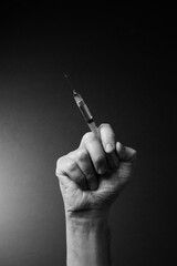 Black and white image of man's clenched hand holding syringe upwards with liquid droplet on needle point, with dramatic lighting
