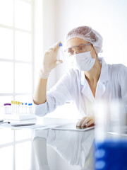 Professional female scientist in protective eyeglasses researching tube with reagents in sunny laboratory. Medicine and science researching