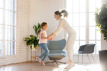 Happy young Caucasian mother and little 7s daughter dance and jump in cozy own living room. Overjoyed mom or nanny have fun engaged in funny game with small girl child in modern renovated home.
