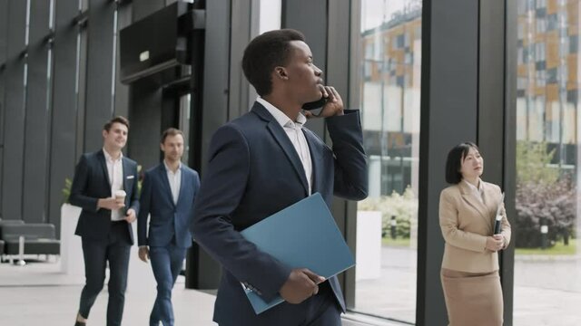 Medium shot of young good-looking African businessman wearing elegant blue suit going along office hall and having telephone chat with colleague