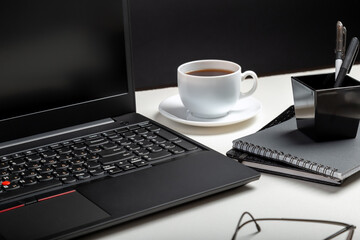 Black laptop on white table black background. Glasses, cup of coffee, office supplies. Home workspace for work or study in Office interior