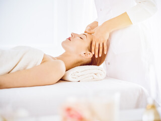 Fototapeta na wymiar Beautiful woman enjoying facial massage with closed eyes in sunny spa center