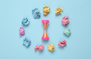 Hourglass and crumpled balls of colored paper on a blue background. Lack of ideas concept