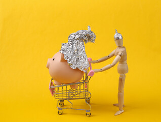Wooden puppet rolls shopping trolley with Piggy bank in foil hat on yellow background