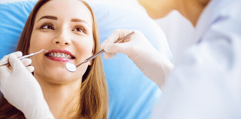 Smiling young woman with orthodontic brackets examined by dentist in sunny dental clinic. Healthy teeth and medicine concept