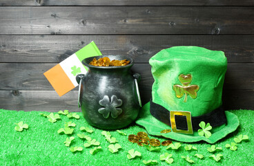 Cast iron pot with treasure, irish flag with clover leaf and leprehaun hat, clover leaves and coins on green grass, dark wooden planks background. Saint Patricks Day banner, poster, flyer, invitation