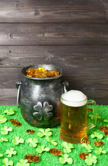 Cast iron pot, decorated by four-petal lucky shamrock leaf, full of leprechaun gold treasure, mug of beer, clover leaves and gold coins on green grass, dark wooden planks background.