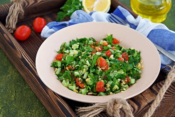 Tabbouleh, parsley, tomato and cucumber vegetable salad with bulgur in a clay plate on an olive concrete background. Salad recipes. Healthly food.