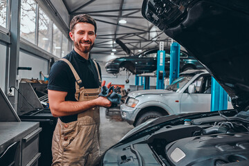 After the work is done. A confident young man in uniform wiping his hands with a rag and smiling while standing in the workshop with a car on the background.