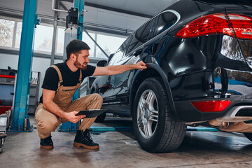 Handsome mechanic in uniform is making notes examining car in auto service.