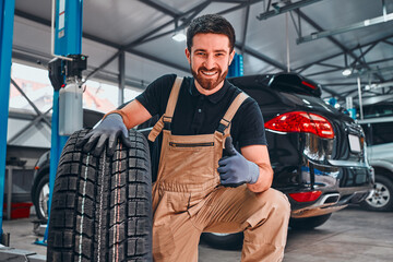 Young smiling mechanic showing thumbs up with car tire.