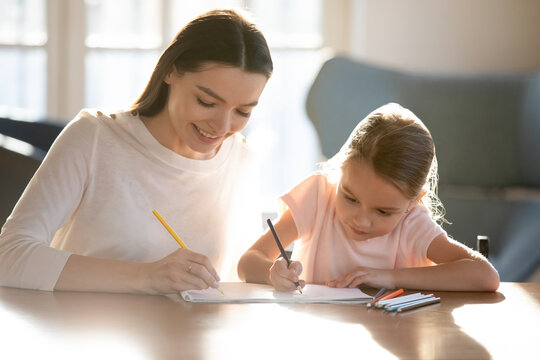 Caring young Caucasian mother and small 7s daughter sit at table at home paint in album together. Smiling loving mom and little girl child have fun drawing on family weekend. Hobby concept.