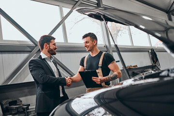 Handsome businessman and auto service mechanic are discussing the work and shaking hands.