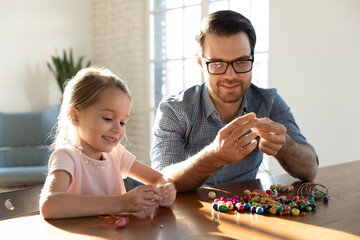 Smiling loving young dad and little daughter have fun make bracelets with colorful beads at home...