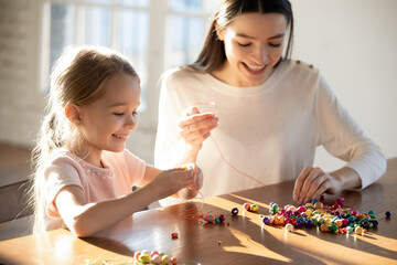 Happy caring young Caucasian mother and little 6s daughter have fun together making bracelets...