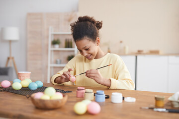 Minimal portrait of smiling African-American girl hand painting Easter eggs with pastel colors while enjoying DIY decorating at home, copy space