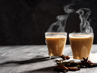 Two mugs of masala tea are on a gray table. Steam comes from them. Hard light, dark background.