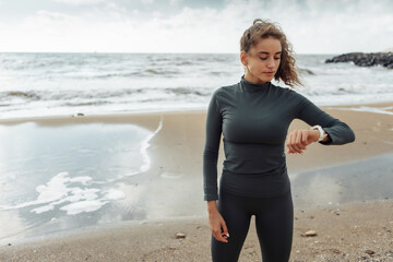 Young purposeful curly-haired fitness woman in sportswear looking at watch on beach