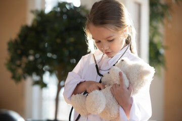 Cute little Caucasian girl kid in medical uniform act as doctor cure examine fluffy toy animal. Happy small 7s child have fun play act as doctor with stuffed teddy bear. Future career concept. - obrazy, fototapety, plakaty