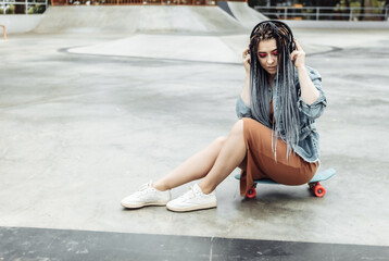 Fototapeta premium Extraordinary female millennial skater with colorful makeup, African braids sits on penny board and listen music in headphones at skate park. Youth, lifestyle concept