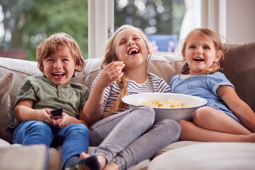 Three Children Sitting On Sofa At Home Laughing And Watching TV With Popcorn